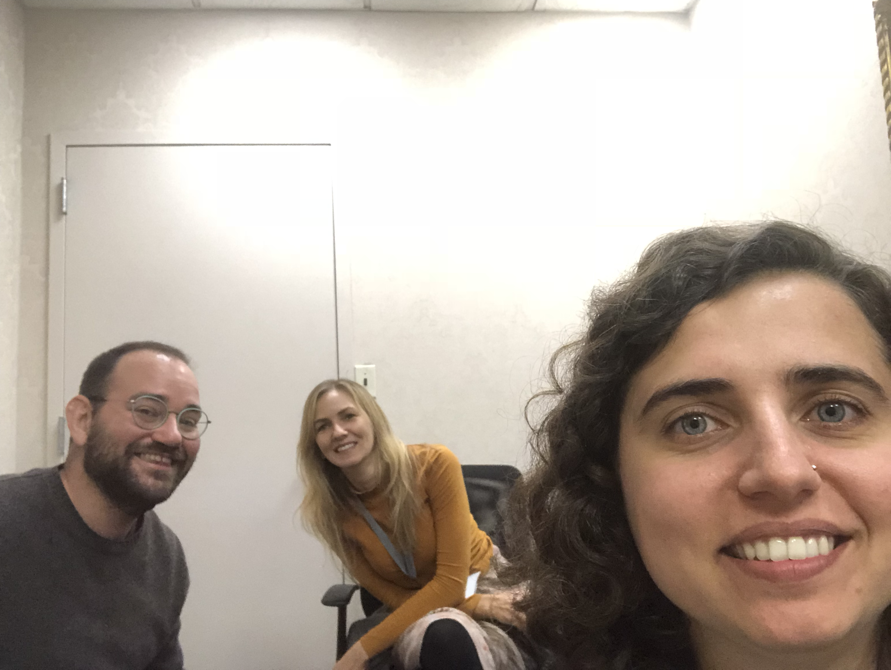 Selfie of three people smiling. From left to right there is a man with flasses and a brown shirt, then a woman with blonde hair and a mustard colored sweater and on the right is a woman taking the selfie.